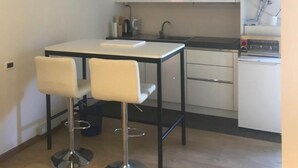 Kompakti keittiö, jossa on valkoiset kaapit, pieni ruokapöytä kahdella tuolilla ja sisäänrakennettu vedenlämmitin.