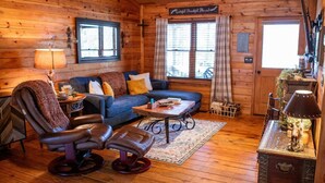 Ein gemütliches Blockhaus-Interieur mit Holzboden, einem blauen Sofa, Ledersesseln, einem kleinen Tisch und einer Lampe.