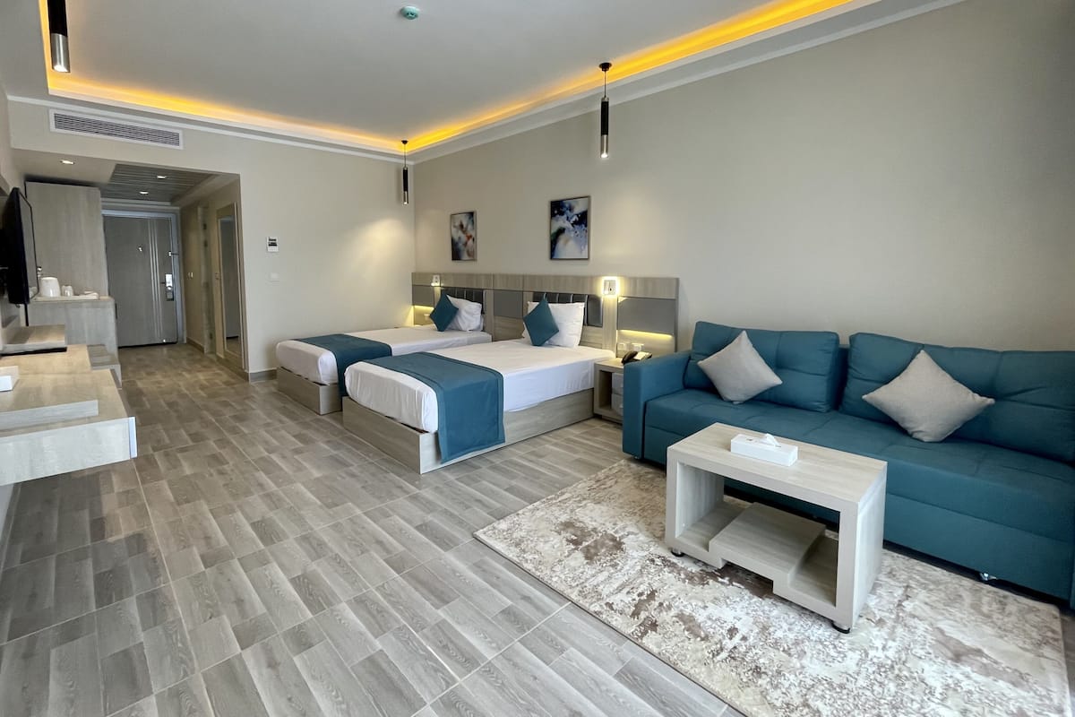 A modern hotel room with two beds, a sofa, a coffee table, and a bathroom visible in the background.
