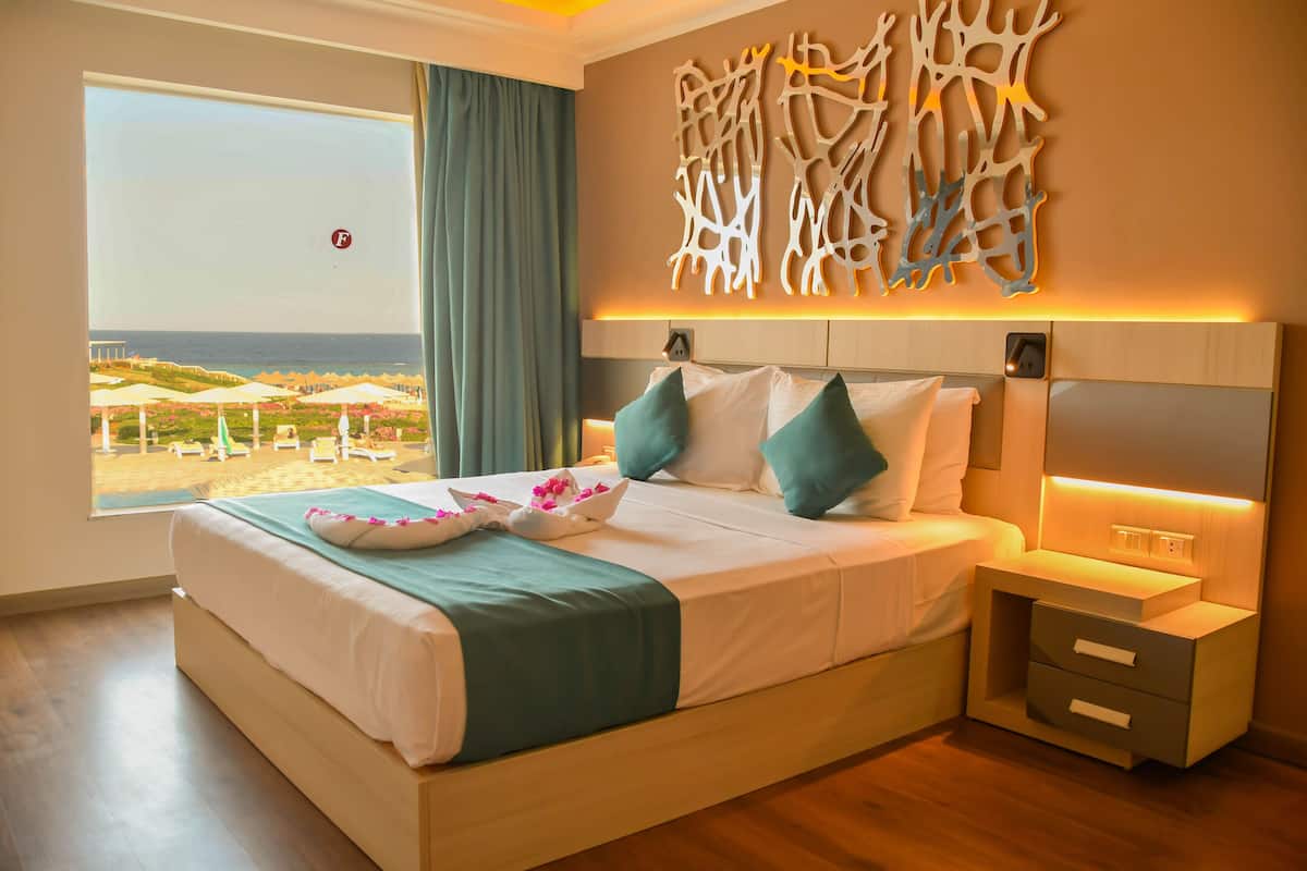 A bedroom with a large bed, a nightstand, a wall-mounted light, and a view of the beach through a window.