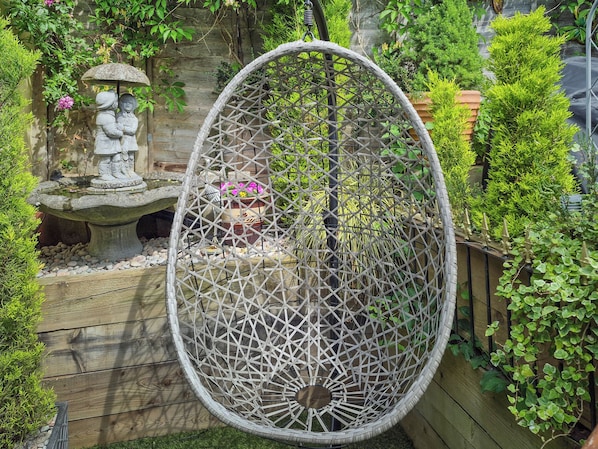 A garden with a decorative stone statue, a wicker swing, and a variety of plants.