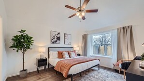 Un dormitorio con cama, ventilador de techo, ventana con cortinas, mesita de noche con lámpara y una planta en maceta.