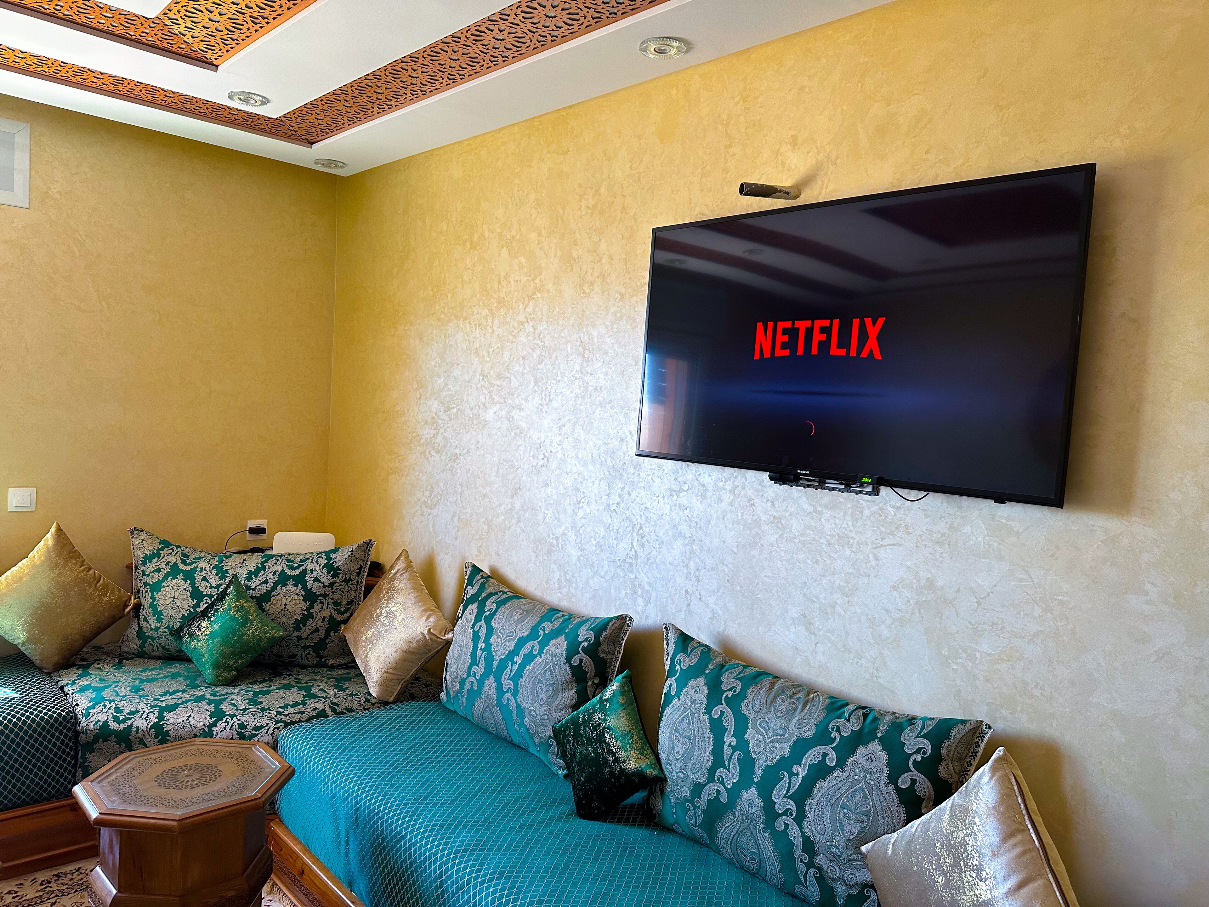 Ein Wohnzimmer mit einer Couch, dekorativen Kissen, einem kleinen Holztisch und einem an der Wand montierten Fernseher, der das Netflix-Logo zeigt.