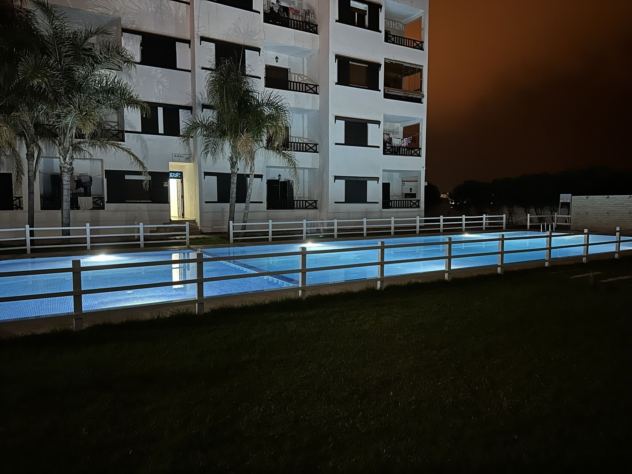 Ein Swimmingpool mit weißem Zaun, ein Gebäude mit mehreren Balkonen und Palmen.