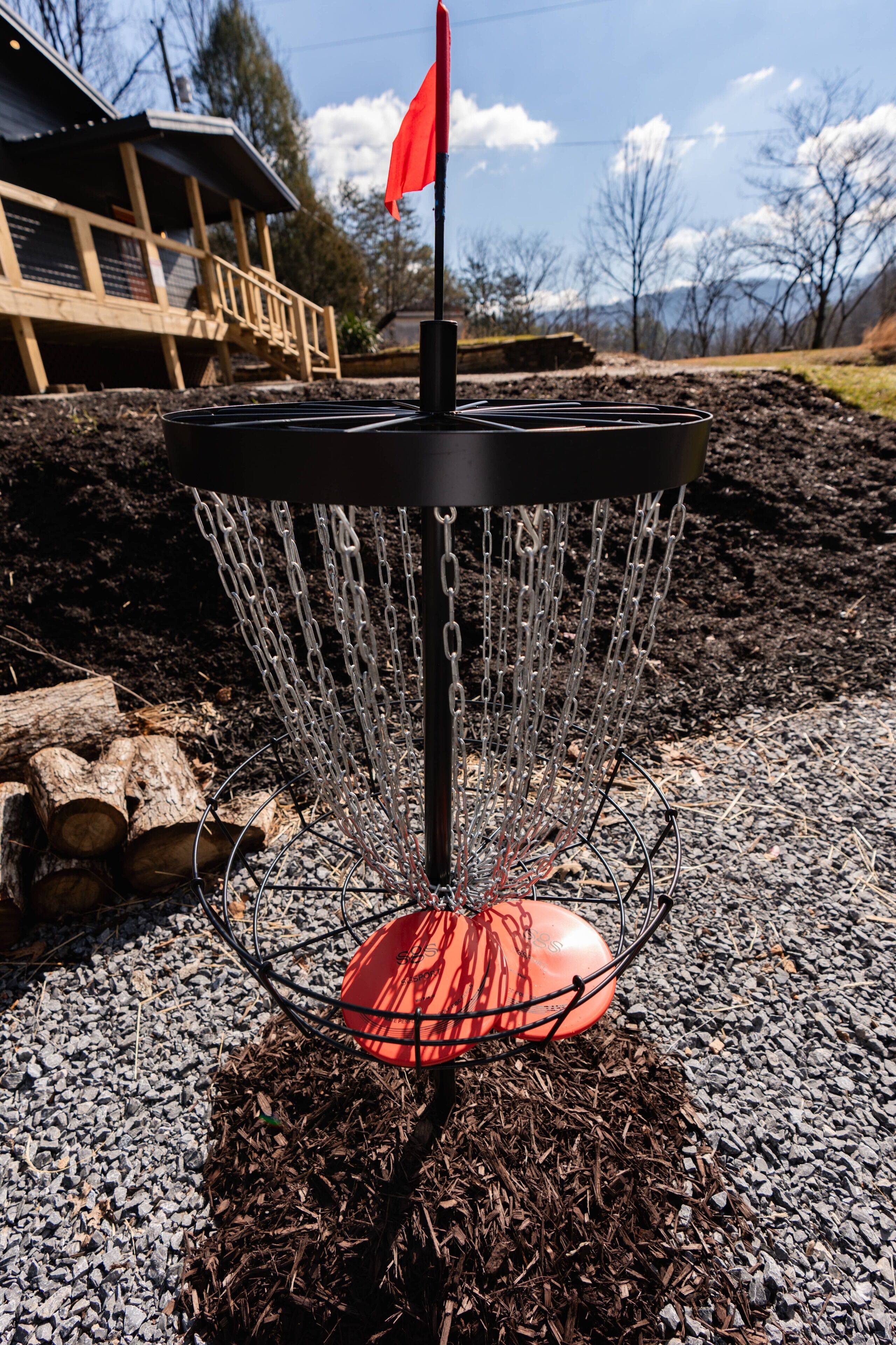 A disc golf basket with a red flag on top, surrounded by gravel and mulch, with a wooden structure and trees in the background.