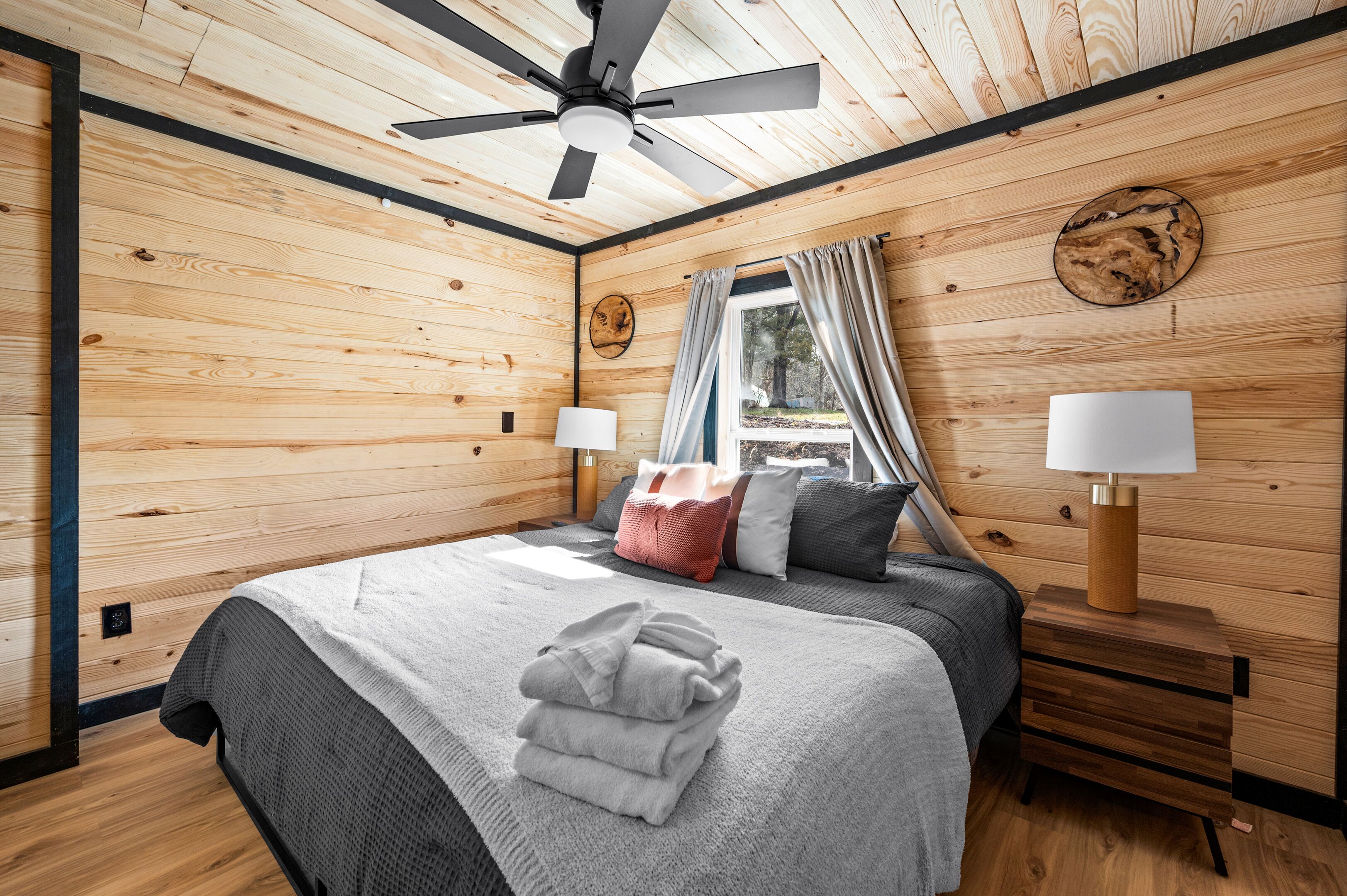 A wooden-paneled bedroom with a bed, two bedside lamps, a ceiling fan, and a window with curtains.