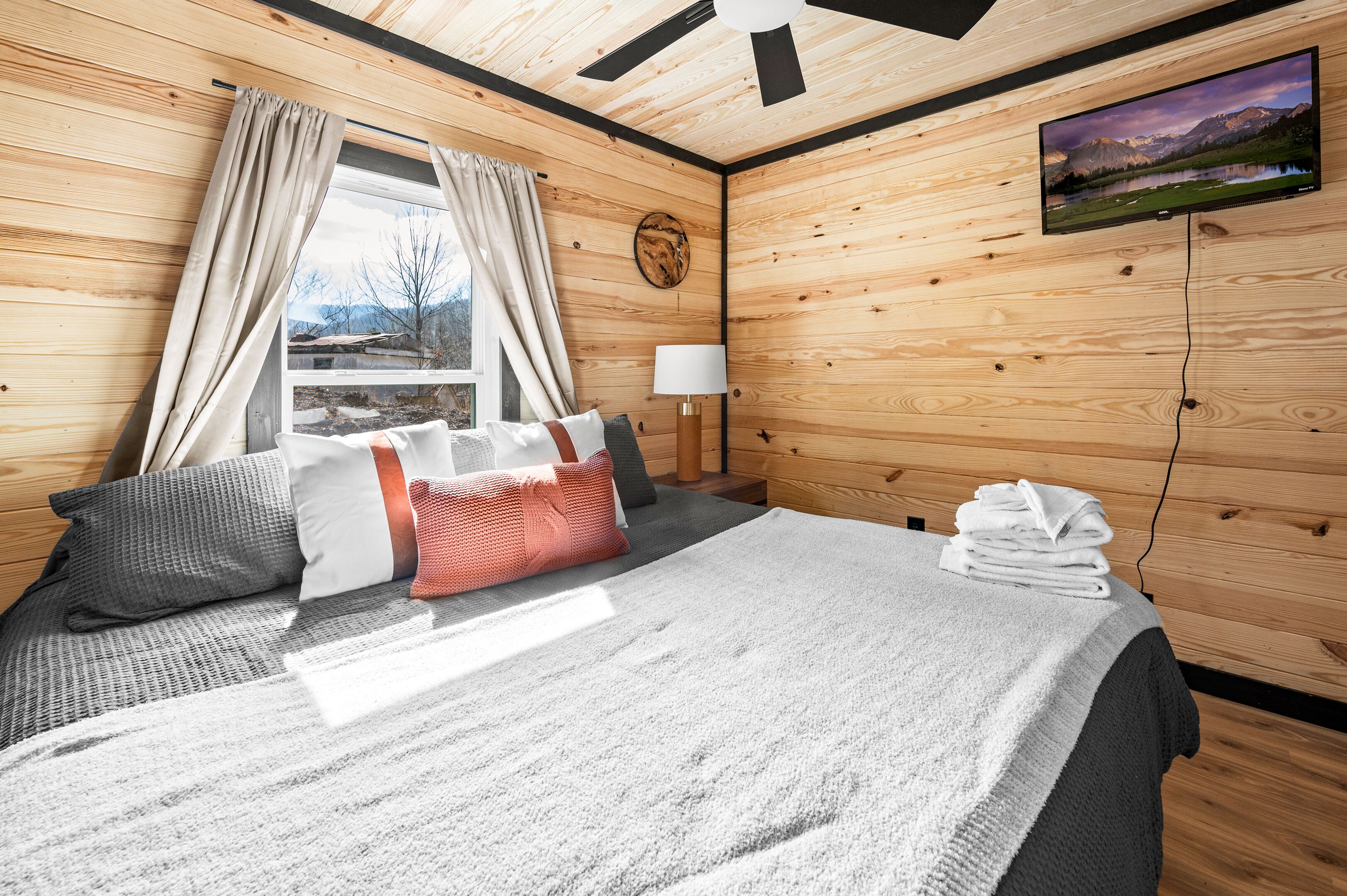 A wooden-paneled bedroom with a bed, pillows, a lamp, a TV, and a window with curtains.