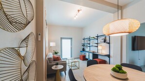 A modern living room with a sofa, coffee table, and dining table. There is a television on the wall and a bookshelf with framed pictures.
