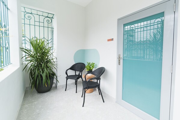 A narrow balcony with two wicker chairs, a large potted plant, and a teal-framed door.