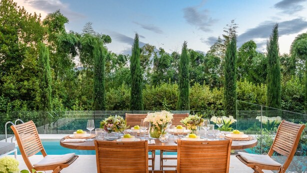 A round dining table with six wooden chairs, set for a meal with white flowers and lemons.