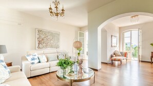 A spacious living room with a sofa, a coffee table with plants, a chandelier, and a large window.