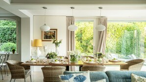 A dining area with a long table set for four, surrounded by wicker chairs, and large windows with curtains.