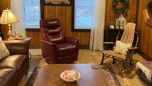 Un salon chaleureux avec un fauteuil en cuir, un fauteuil à bascule en bois, une table basse sur laquelle se trouve un panier de fruits, et un tableau représentant un paysage, accroché au mur.