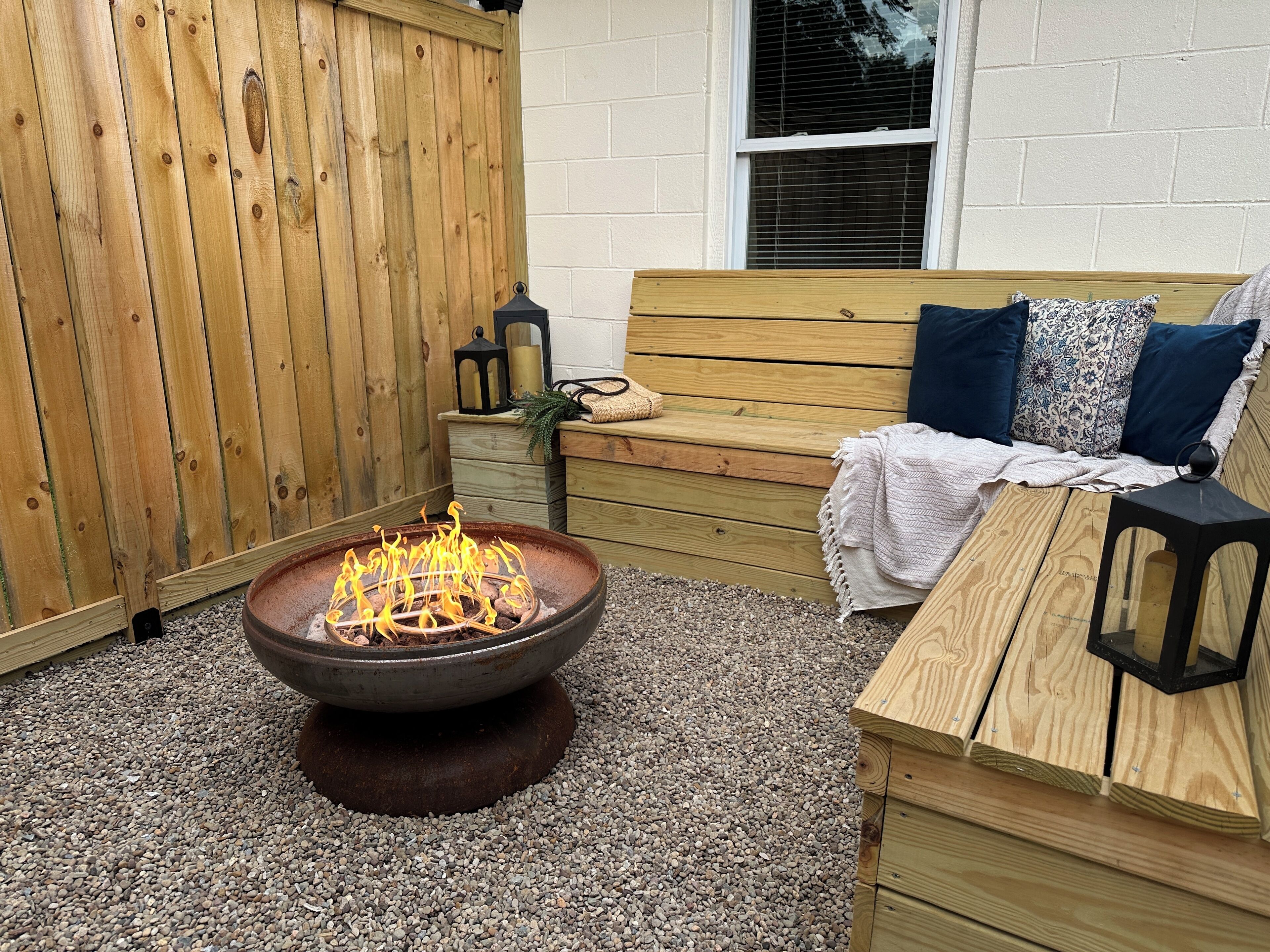 A cozy outdoor seating area with a fire pit, wooden furniture, and a white brick wall in the background.