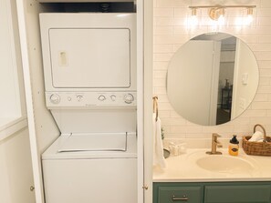 Ein Waschraum mit einer Frontlader-Waschmaschine und einem Trockner, einem Waschbecken mit Spiegel darüber und einem Korbwäscheschrank auf der Arbeitsfläche.