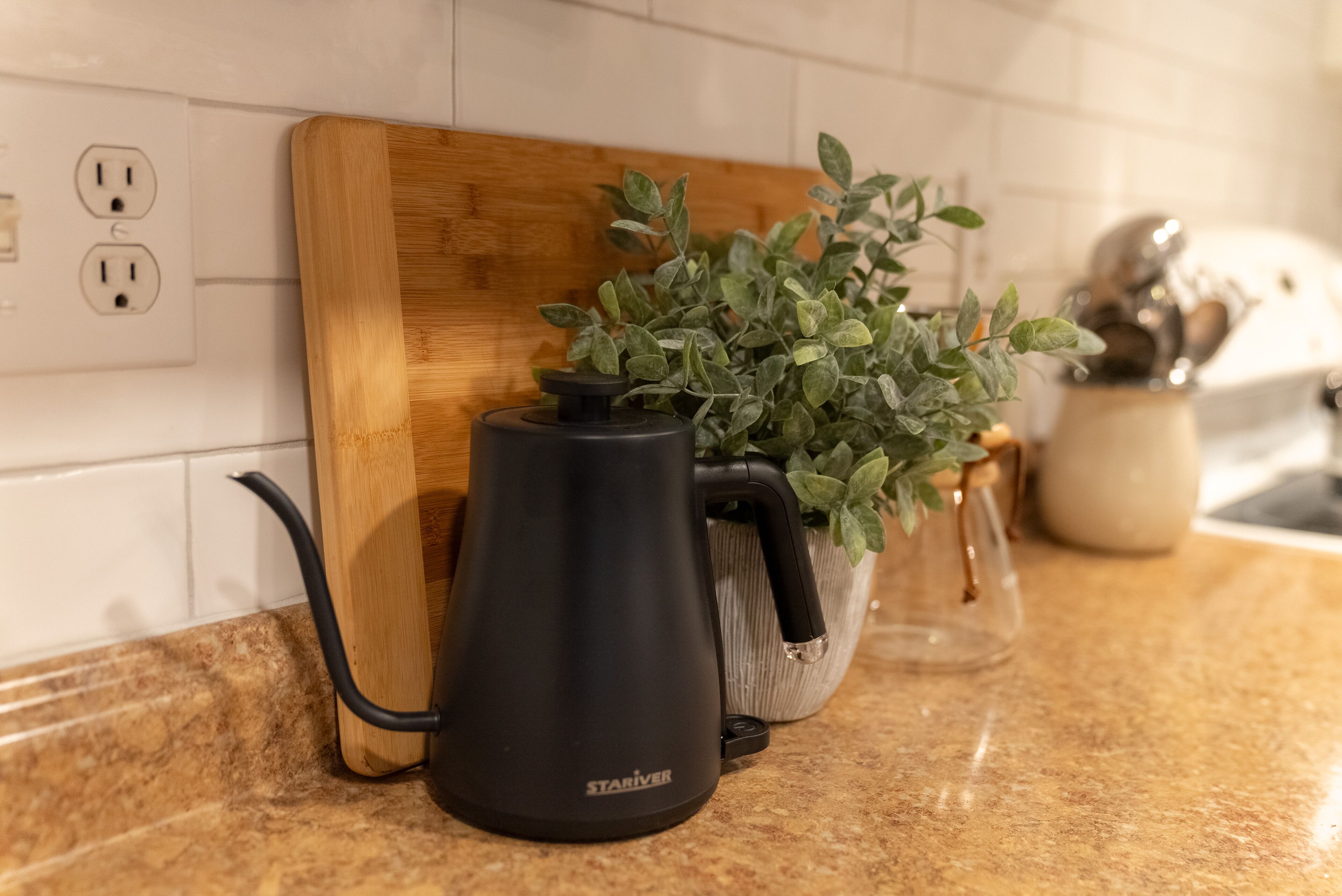 Een keukentafel met een zwarte elektrische waterkoker, een houten snijplank en een potplant.