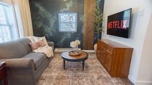 Un salon avec un canapé gris, une table basse ronde surmontée d’un vase, un meuble en bois et un téléviseur fixé au mur qui affiche le logo « NETFLIX ».