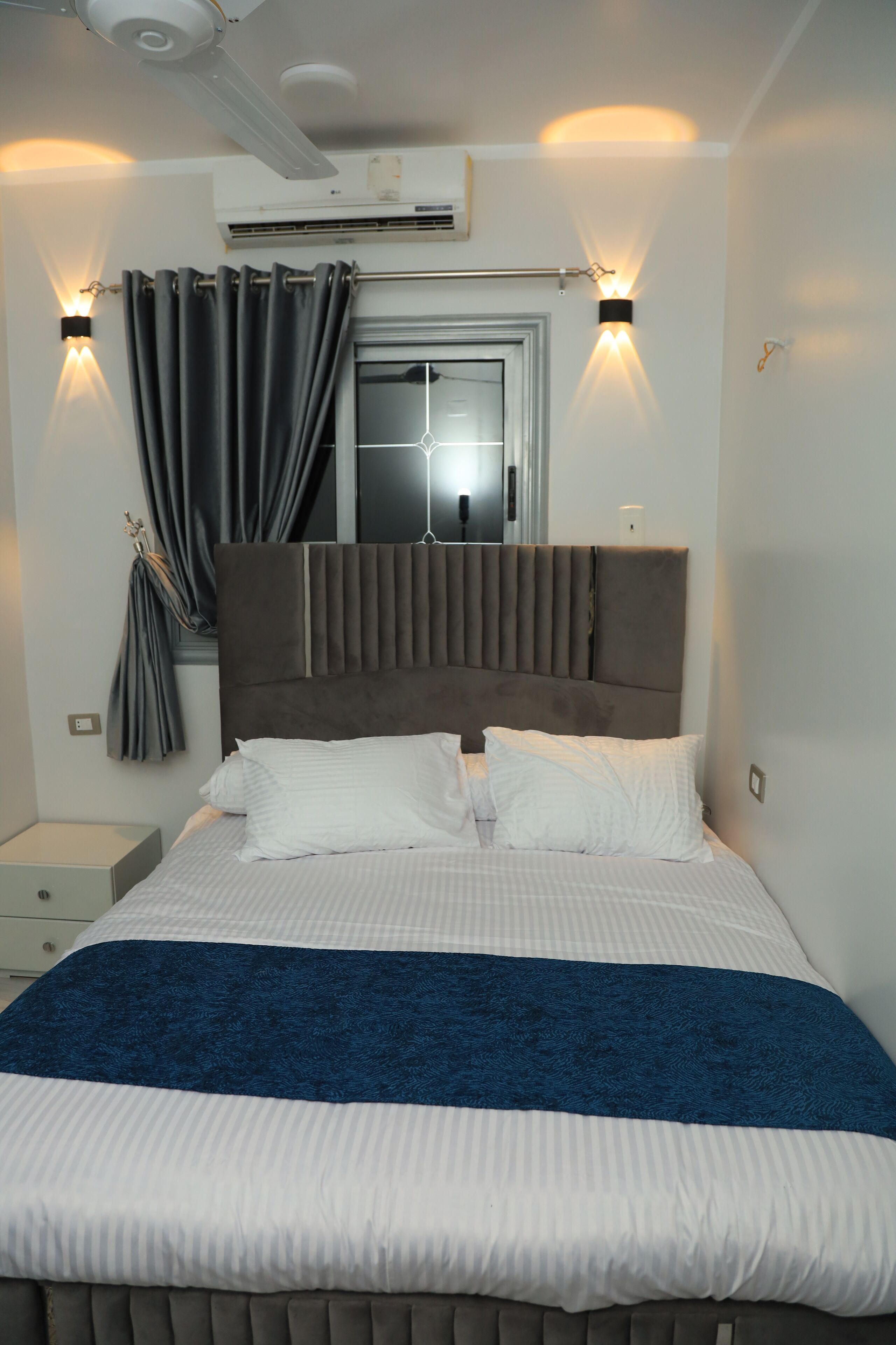 A neatly made bed with a blue blanket, a headboard with vertical slats, and two wall-mounted light fixtures.