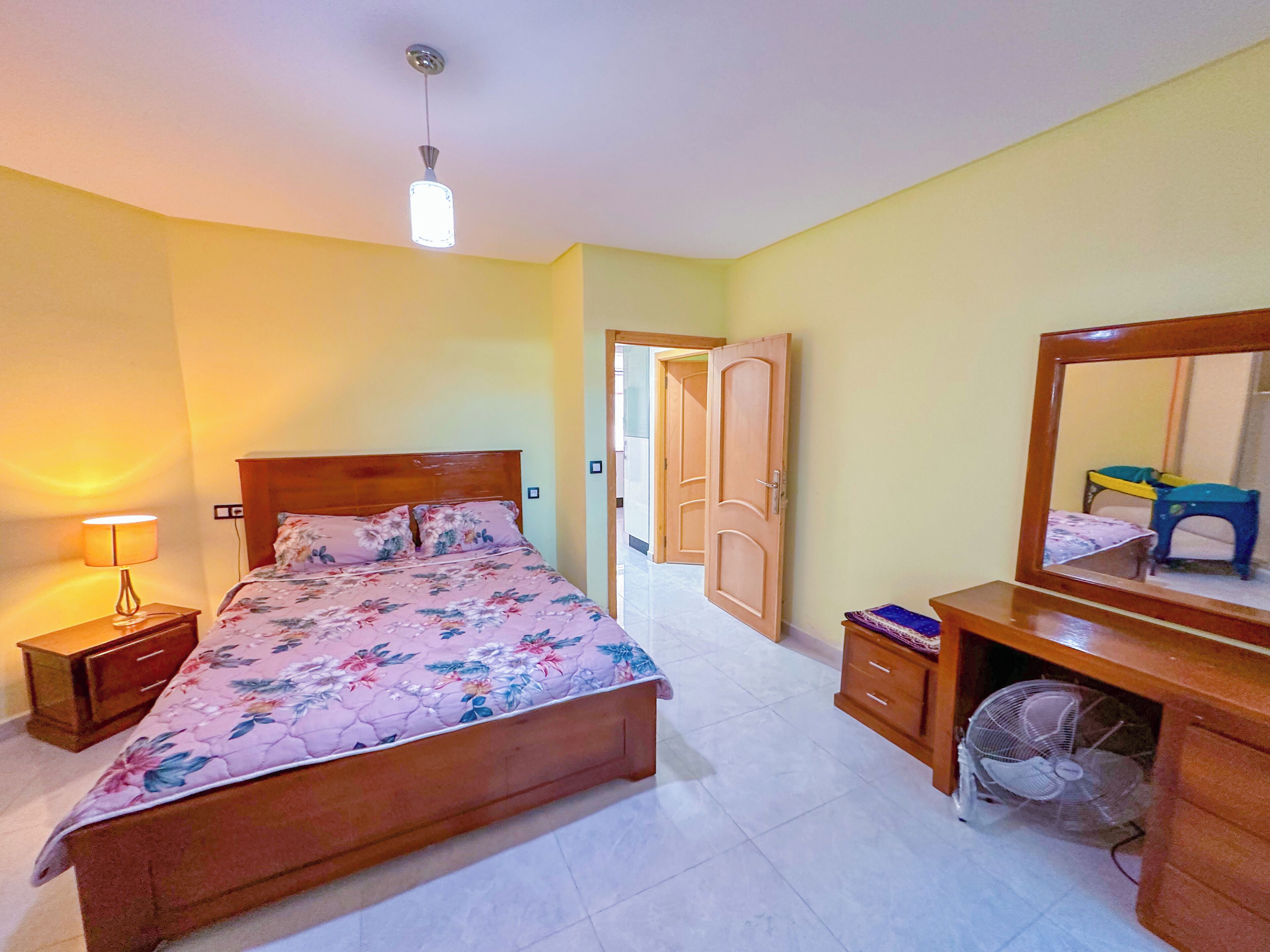 Une chambre à coucher comprenant un lit, une lampe de chevet, une commode en bois, un miroir et une porte donnant sur une autre pièce.