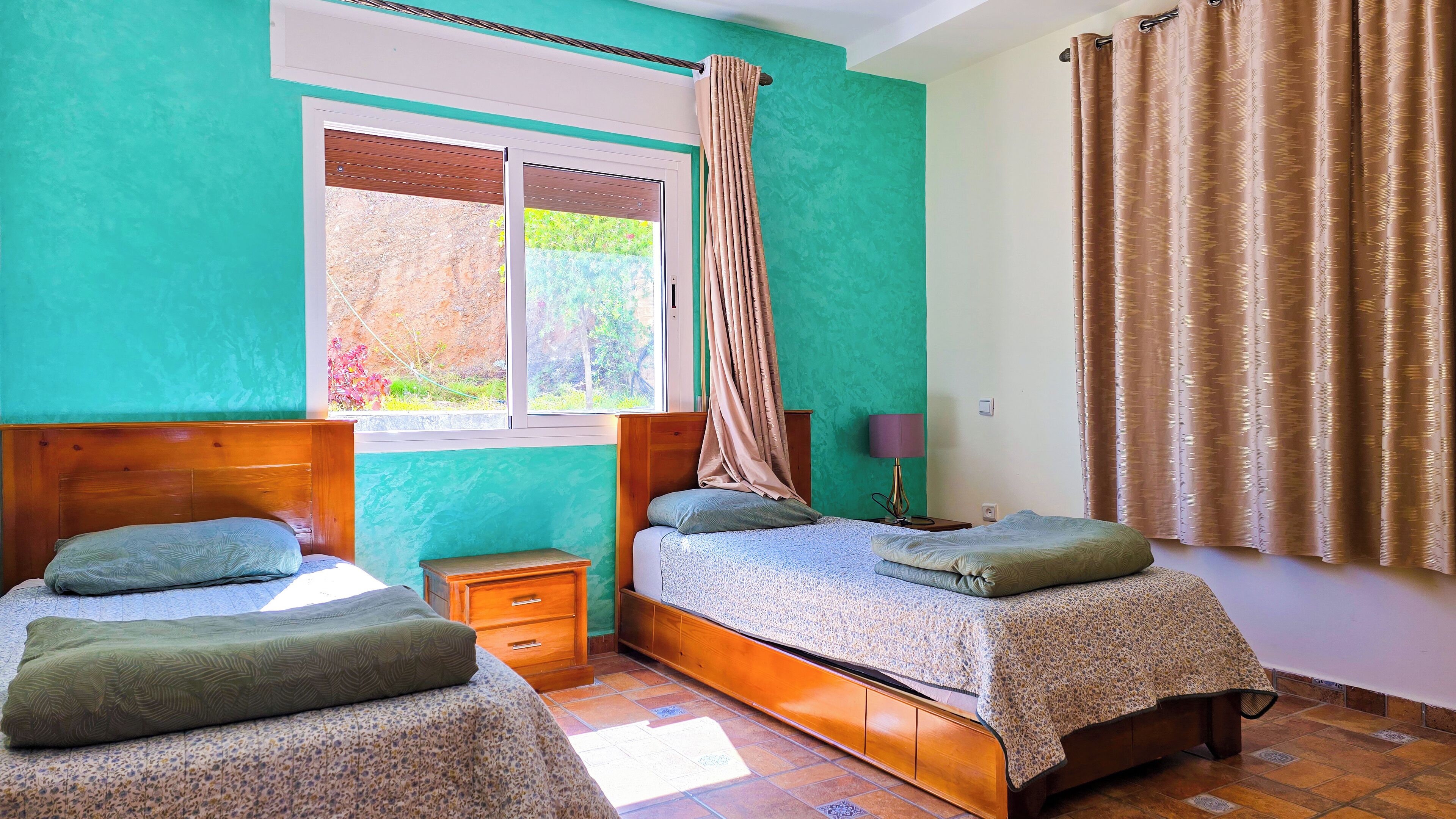 Une chambre avec deux lits, une fenêtre avec des rideaux, une table de chevet avec une lampe, et une vue sur la verdure à l’extérieur.