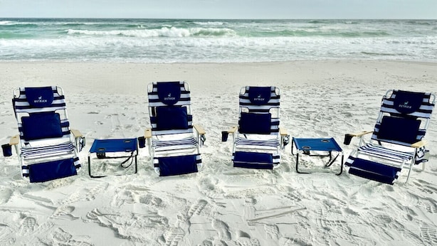 Vier Strandstühle mit blau-weiß gestreiften Kissen und ein blauer Tisch, auf einem sandigen Strand mit sichtbaren Fußabdrücken aufgestellt.