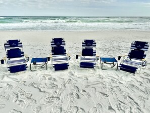 Cuatro sillas de playa con cojines a rayas azules y blancas y una mesa azul, dispuestas sobre una playa arenosa con huellas visibles.