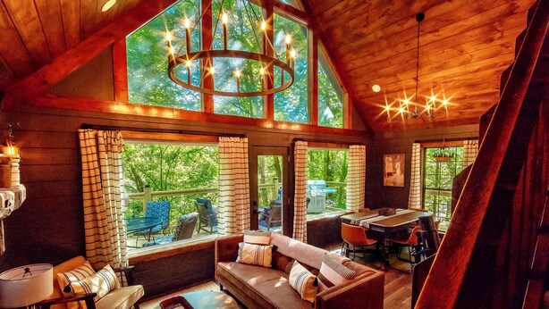 A cozy cabin interior with a wooden ceiling, large windows, a sofa, a dining table, and a piano. - NOT YOUR GRANDMA’S CABIN! King Loft, WIFI, Game Room, Hot Tub & Fire Pit! (Almond)