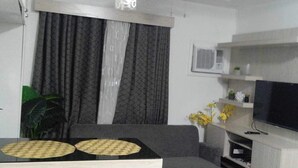 A living room with a sofa, a coffee table with two placemats, a television on a stand, a plant, and a window with curtains. - 1 Br condo T2-8H (Cagayan de Oro)