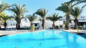 A clear blue swimming pool with a metal ladder, surrounded by palm trees and white buildings with balconies. - Trinity House (Costa Teguise)