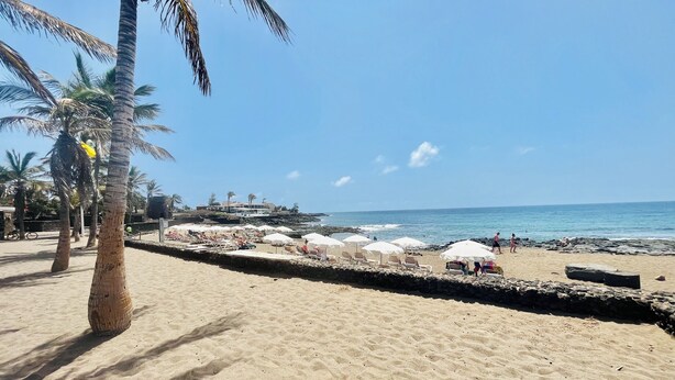 A beach with palm trees, beach chairs, umbrellas, and a rocky shoreline. - Trinity House (Costa Teguise)