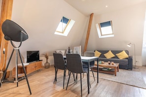 Un salon moderne comprenant un coin repas, un canapé avec des coussins jaunes, un téléviseur et un lampadaire sur trépied.