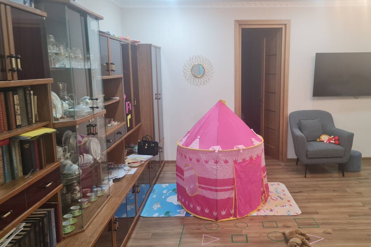 Uma sala de estar com uma estante de madeira, uma tenda infantil rosa, uma poltrona cinza e uma televisão de tela plana fixada na parede.