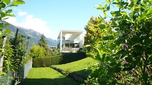 Una casa moderna con fachada blanca, rodeada de exuberante vegetación y un telón de fondo montañoso.