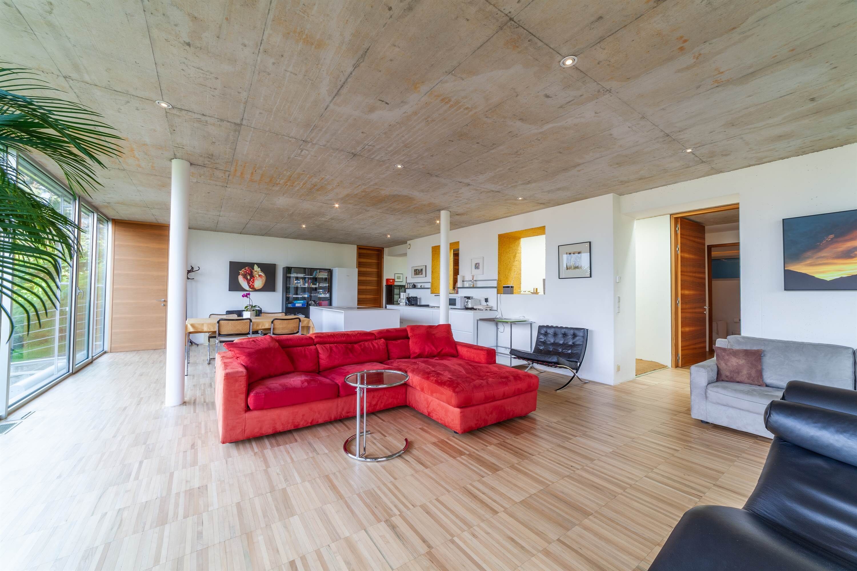 Un salón espacioso con un sofá seccional rojo, un sofá gris y un sillón de cuero negro. Se observa una mesa redonda y un comedor al fondo.
