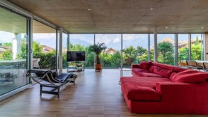 Amplia sala de estar con un sofá rojo, un sillón negro y un televisor sobre un mueble bajo. Amplias ventanas ofrecen vistas a zonas verdes y edificios.