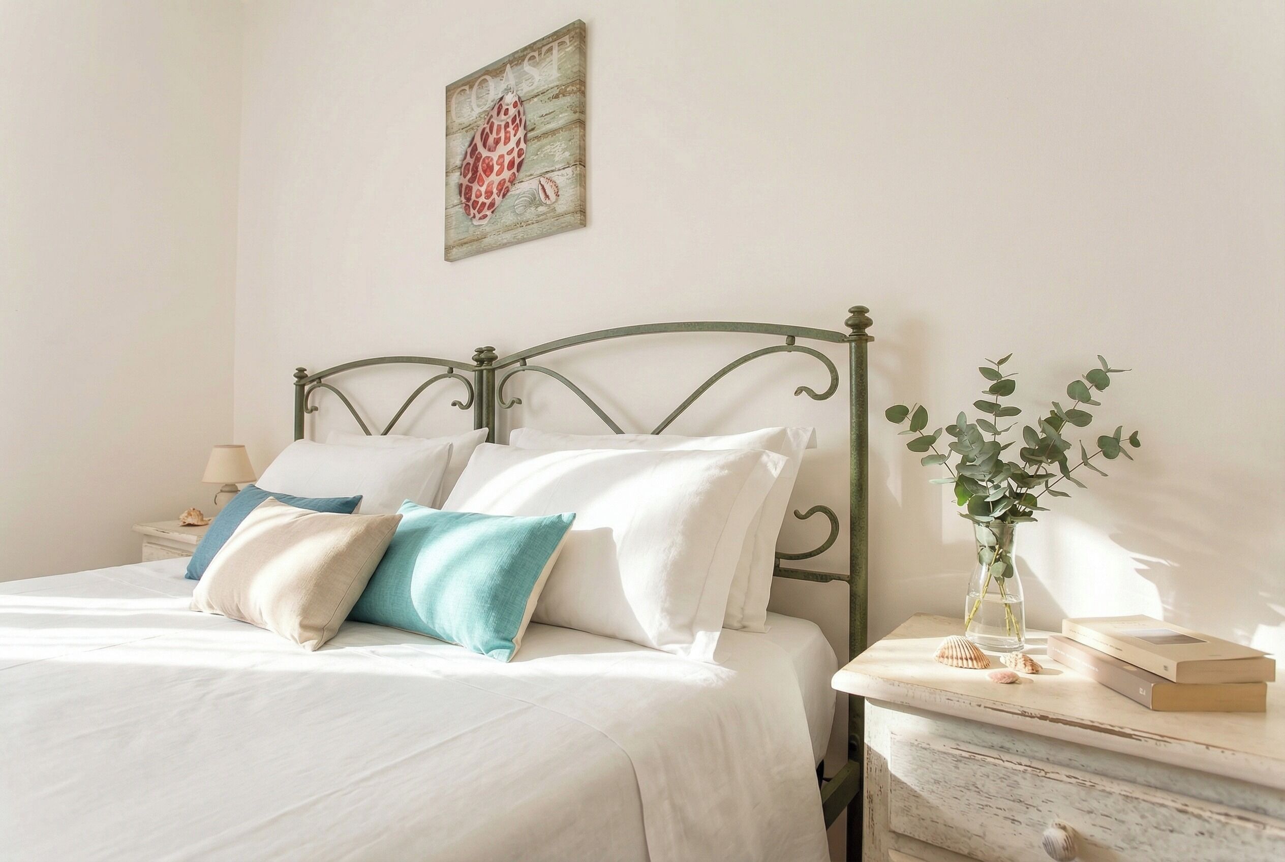 Un lit bien fait, avec des oreillers blancs et turquoise, une tête de lit en bois, une table de chevet ornée d’un vase contenant des feuilles vertes et un tableau encadré accroché au mur.