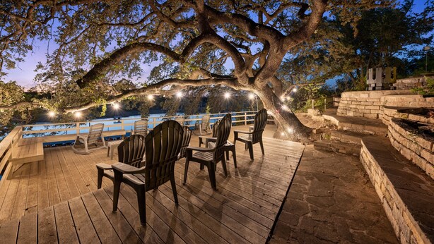 Una terraza de madera con sillas y una mesa, con vista a un cuerpo de agua, adornada con luces colgantes y una gran sombra de árbol.