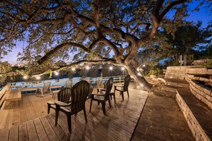 A wooden deck with chairs and a table, overlooking a body of water, with string lights and a large tree providing shade.