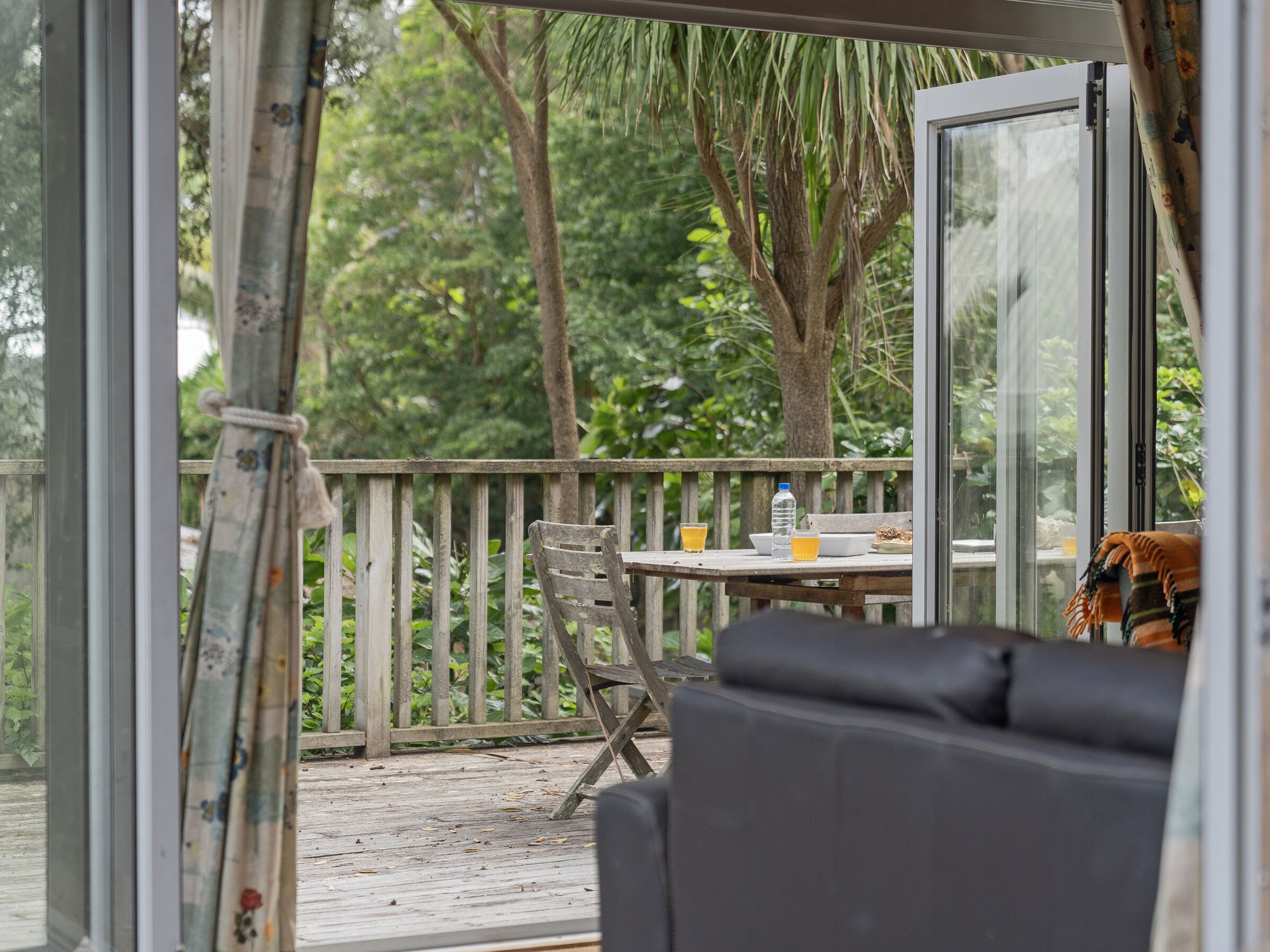Una terraza de madera con una mesa, sillas, un banco y un ventanal que da a un jardín exuberante.