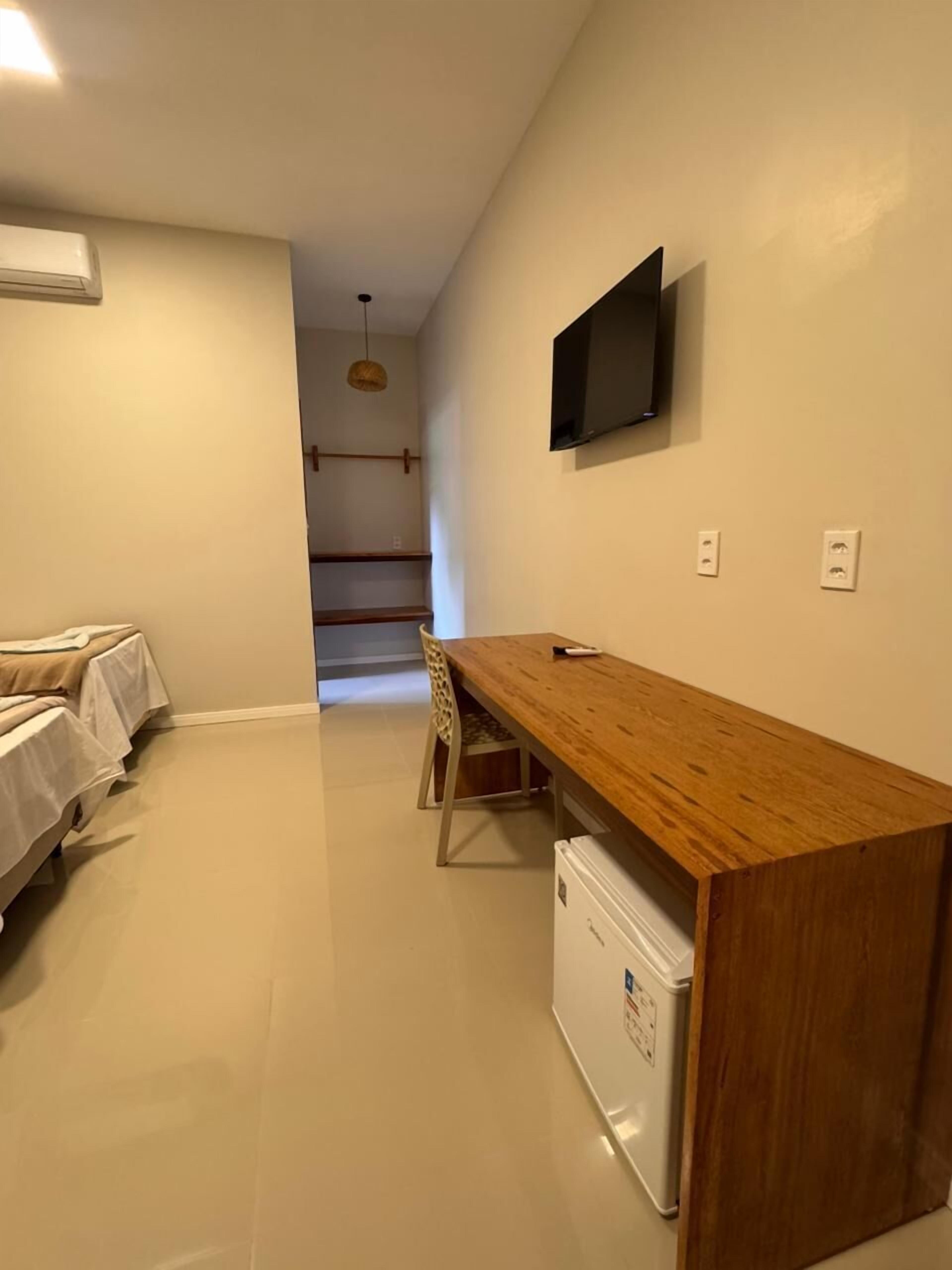 A small, clean room with a wooden desk, a chair, a bed, a TV mounted on the wall, and a small refrigerator.