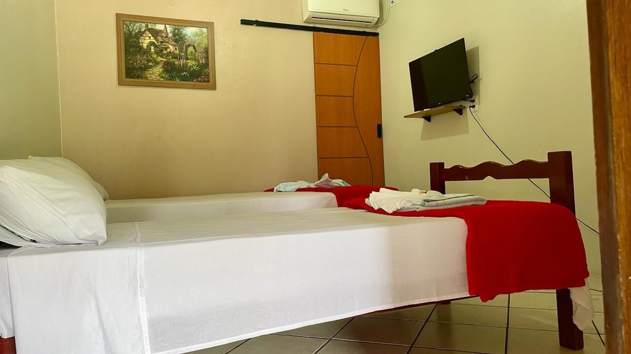A single bed with white bedding and a red blanket, a wooden headboard, a mounted television, and a painting on the wall.