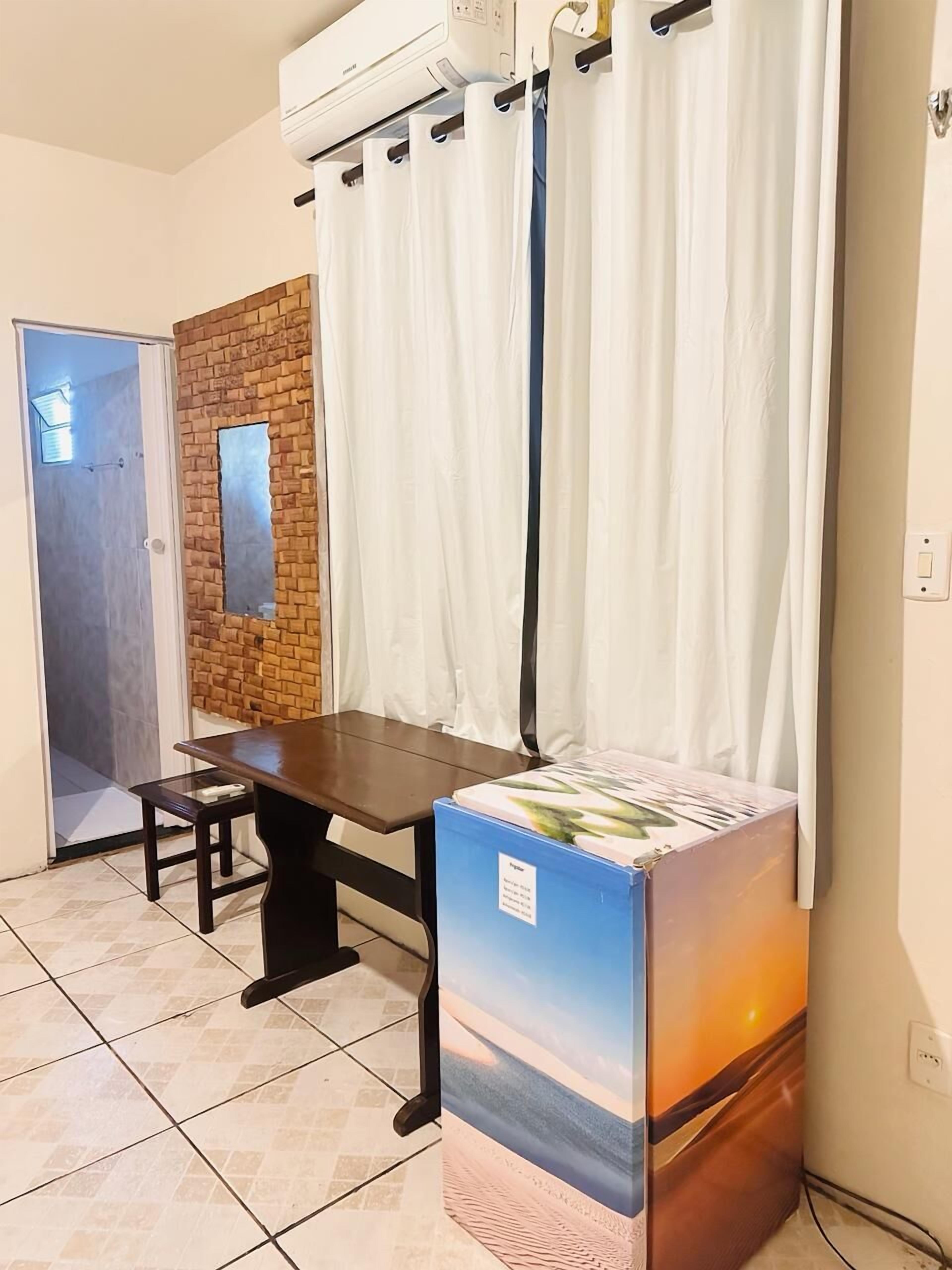 Una habitación con piso de baldosas, una mesa de madera, un banco, aire acondicionado instalado en la pared, una puerta y una ventana con cortinas.