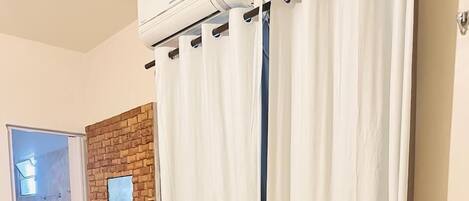 Una habitación con piso de baldosas, una mesa de madera, un banco, aire acondicionado instalado en la pared, una puerta y una ventana con cortinas.