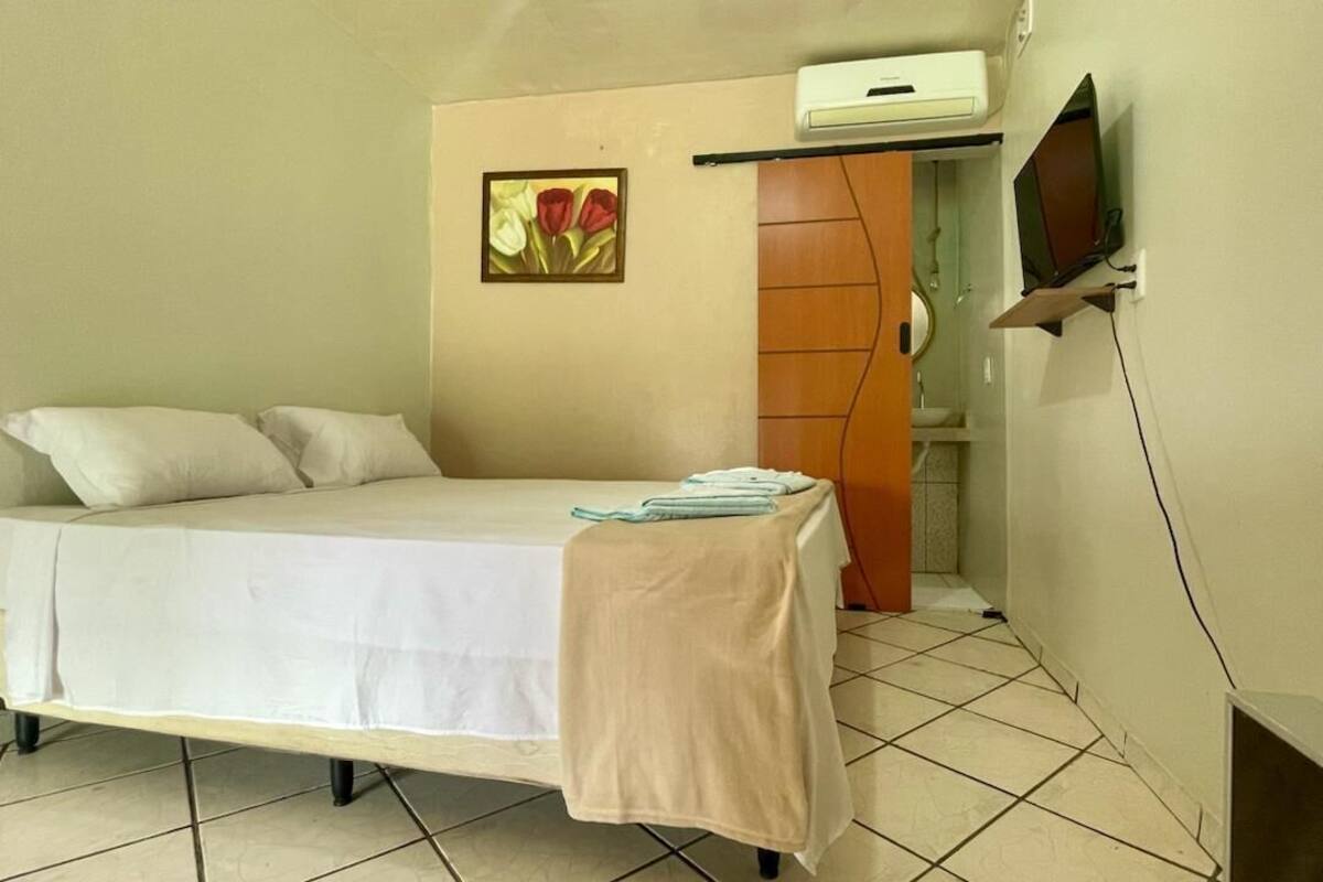 Ein Einzelbett mit weißer Bettdecke und einer beigen Decke, ein kleiner Tisch mit einem Handtuch, ein an der Wand befestigtes Fernsehgerät und eine Klimaanlage.