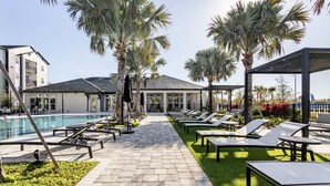 A poolside area with lounge chairs and a covered seating area, surrounded by palm trees and a building with large windows. - Boho Chic Luxury Retreat Minutes to Theme Parks, Restaurants and Entertainment. (Kissimmee)