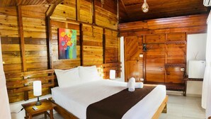 Un dormitorio tipo cabaña de madera con una cama grande, una mesita de noche con lámpara y un cuadro de colores en la pared.