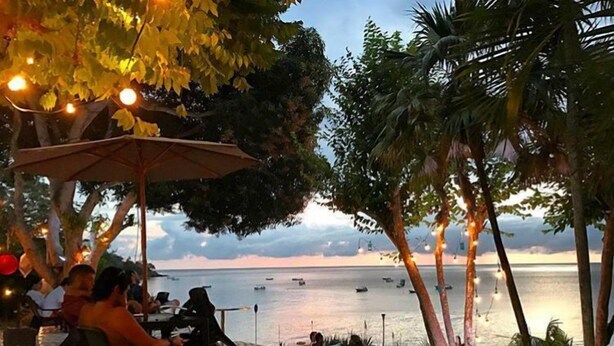 Un espacio al aire libre con vistas al mar, mesas, sillas y luces colgantes.