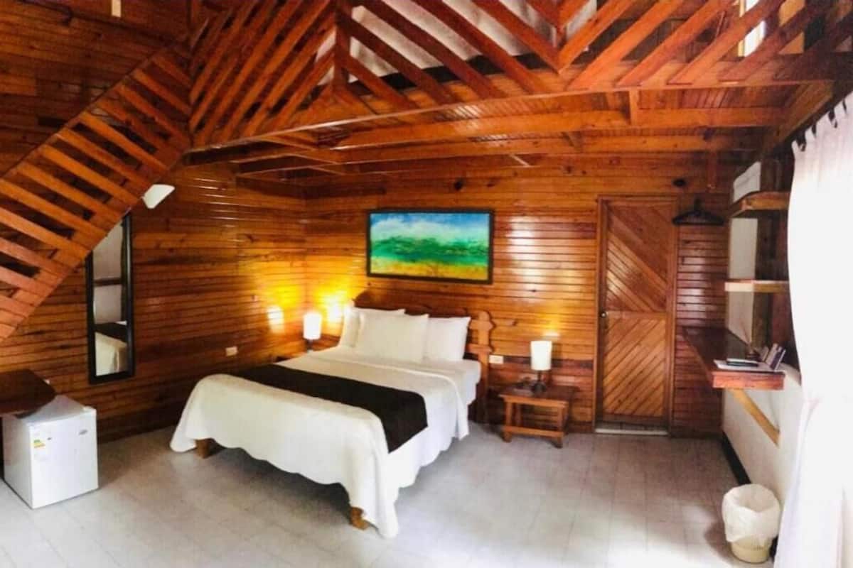 A cozy attic bedroom with a wooden sloped ceiling, a bed with white linens, two bedside lamps, and a painting on the wall.