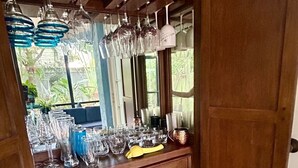 Et område med en træbar, flere glas og flasker, et gult stof og et vindue med grønt udenfor.