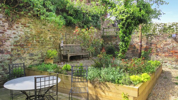 A patio with a round table and two chairs, surrounded by a brick wall and a garden bed with various plants. - 4 bedroom accommodation in Bridlington (Bridlington)